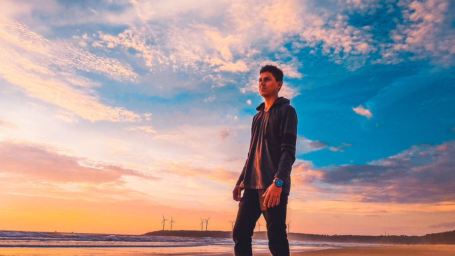 Young man standing at beach against sky during sunset