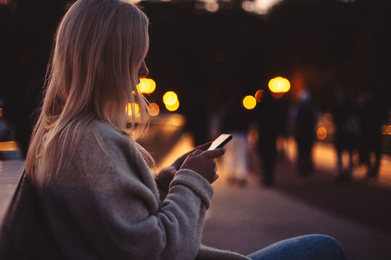 Young woman using smart phone while sitting on bench in city at dusk