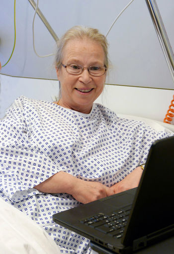 Smiling senior woman looking at laptop while sitting on bed at hospital