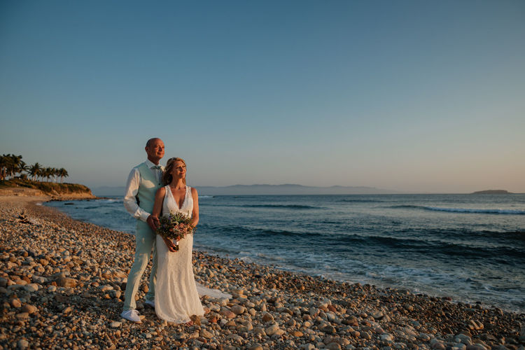 Bride and groom standing on beach against clear sky