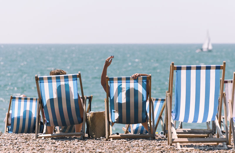 People relaxing on deck chairs at beach against clear sky