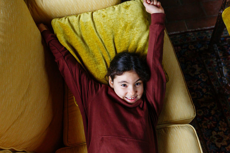 Upper view of smiling girl with dark hair laying on yellow sofa with hands up