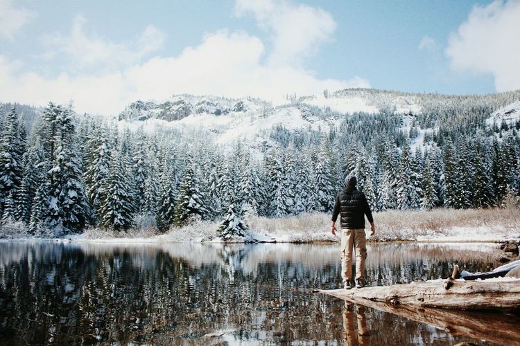 Rear view of man standing at lakeshore against mountain during winter