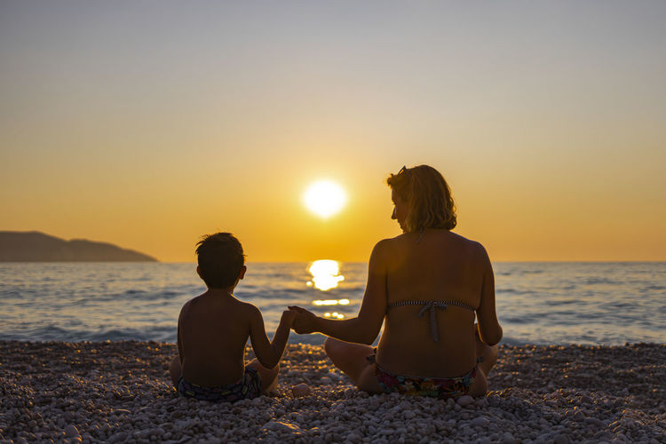 Rear view of mother and son sitting on beach during sunset