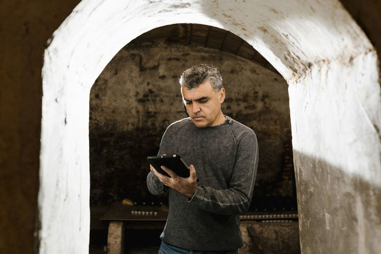 Mature man using digital tablet while standing at archway in cellar
