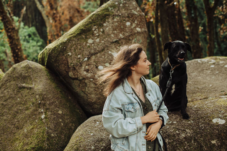 Girl looking at a black dog on a leash sitting on rock in the forest