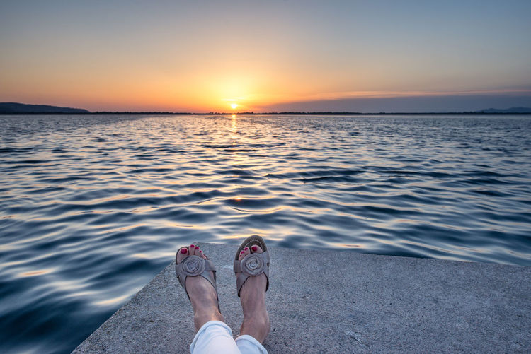 Low section of woman by sea against sky during sunset