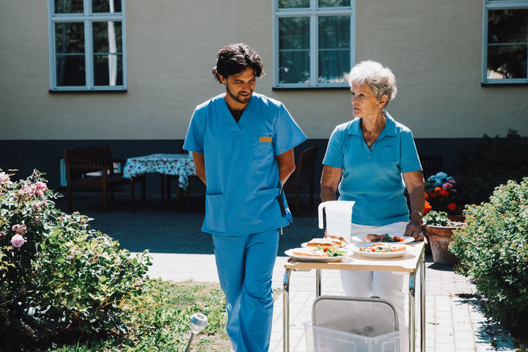 Male nurse walking with senior woman pushing food cart against nursing home