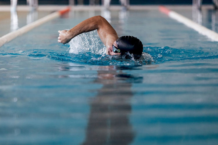 Male swimmer in cap and goggles performing sidestroke while swimming in rippled pool