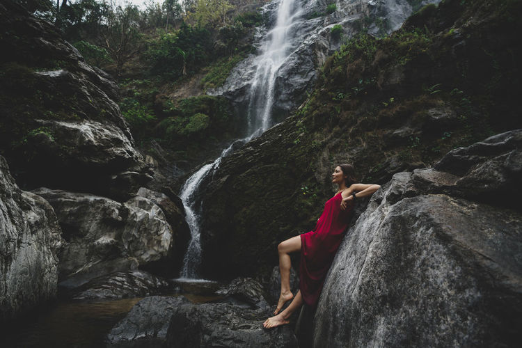 Side view of woman leaning on rock by waterfall