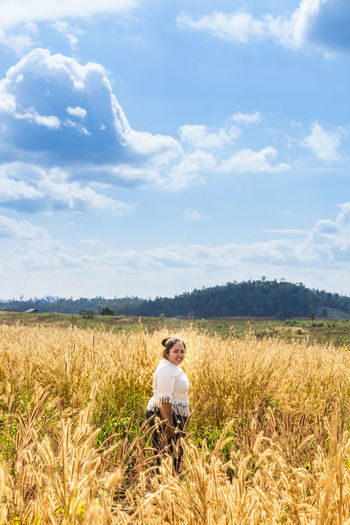 Portrait of young woman standing on grassy field against cloudy sky during sunny day
