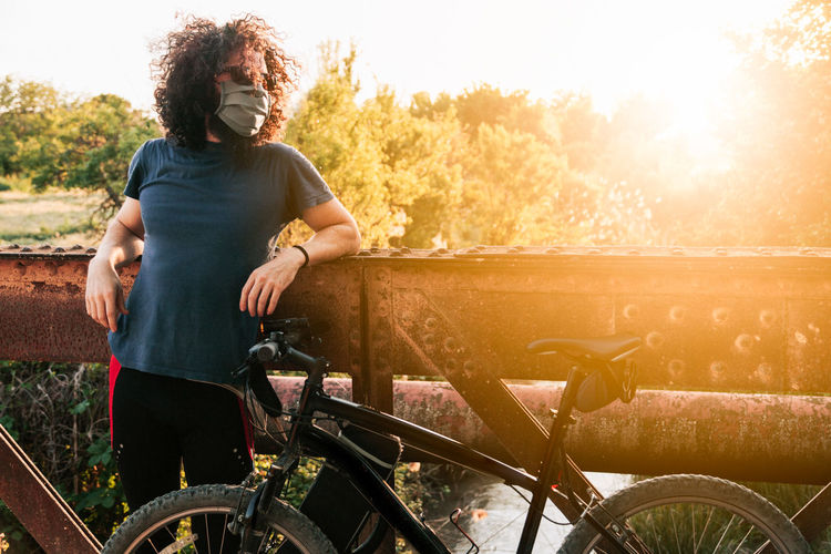 Man wearing mask with bicycle standing by railing at sunset