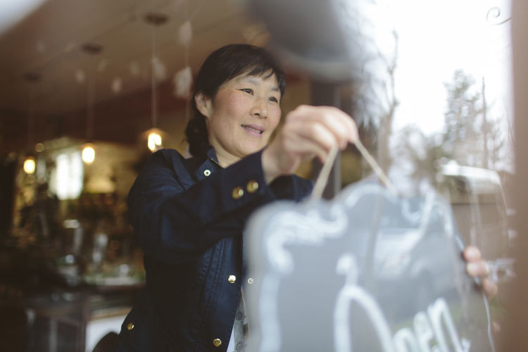 Female owner hanging open sign on window of plant shop seen through glass