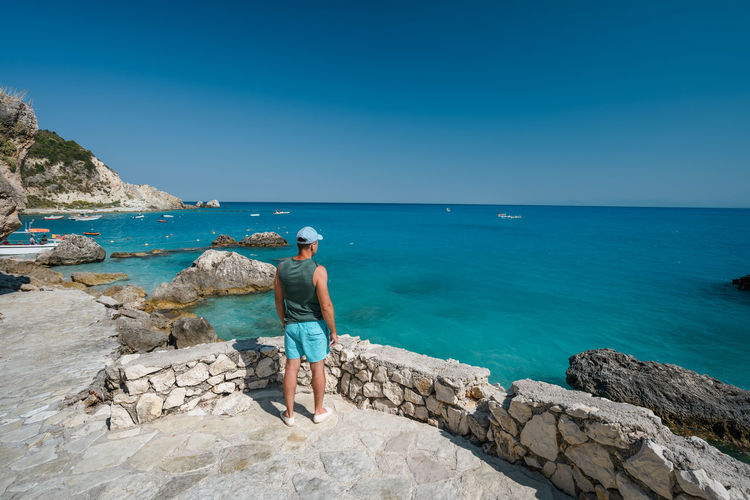 Man standing on rocks by sea against blue sky