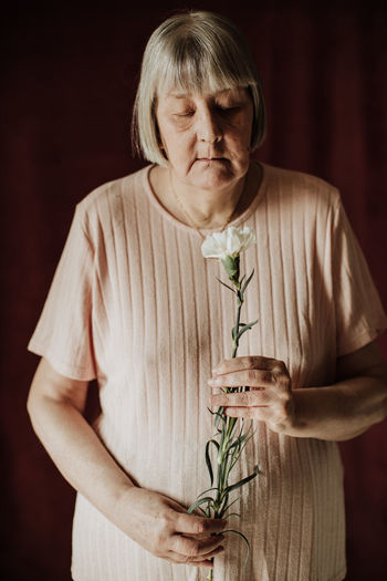 From above thoughtful old female with gray hair with closed eyes holding bouquet of white carnation at home