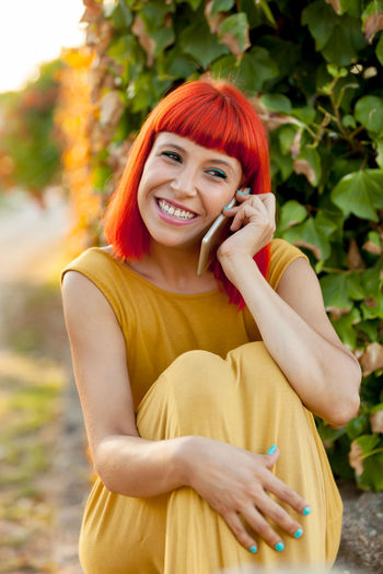 Smiling woman talking on phone while sitting against plant