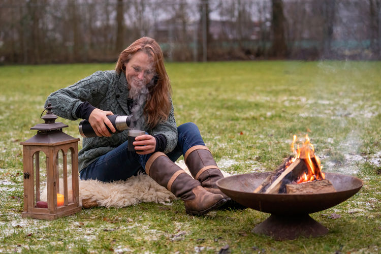  woman is sitting outside on a fur by the campfire in the forest. she pours coffee selective focus.