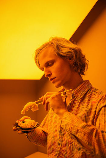 Young man eating food while sitting against wall in room