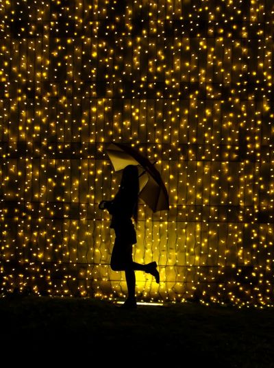 Silhouette woman with umbrella standing against illuminated string lights at night