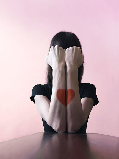 Woman with heart shape on hand covering face against wall