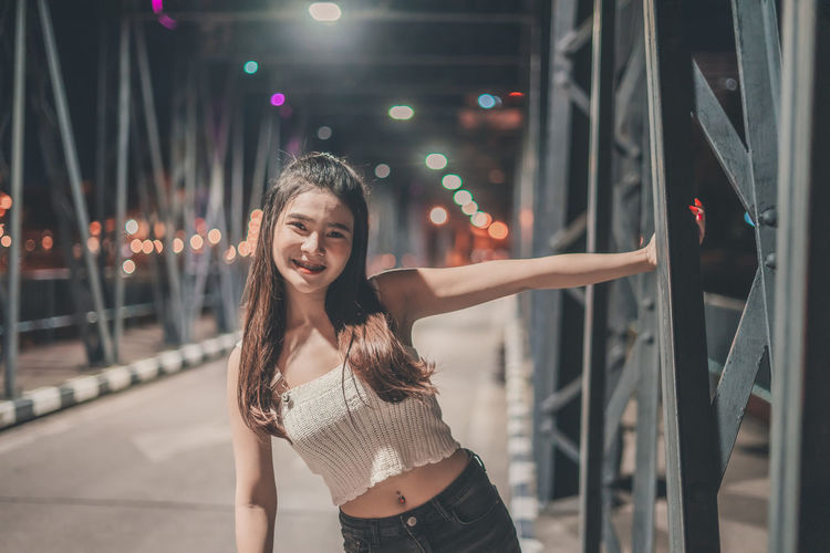 Portrait of smiling young woman standing on footbridge in city at night