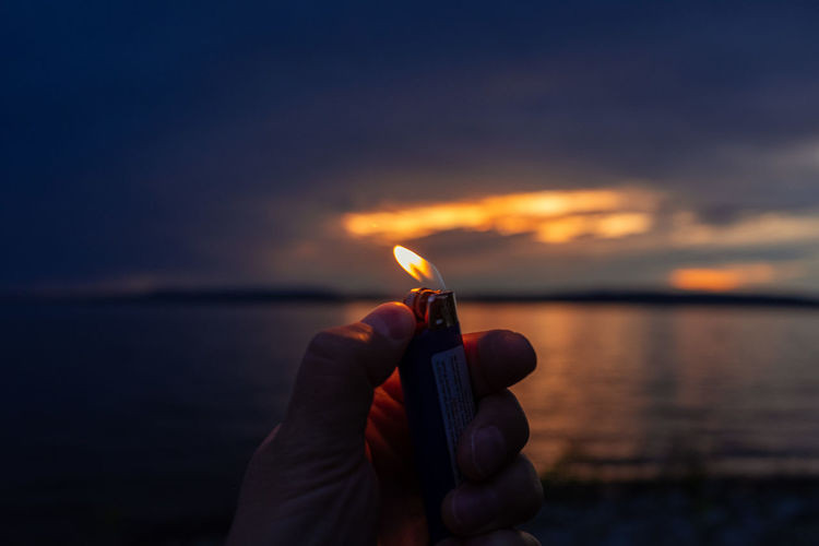 Cropped hand holding illuminated cigarette lighter against sky during sunset