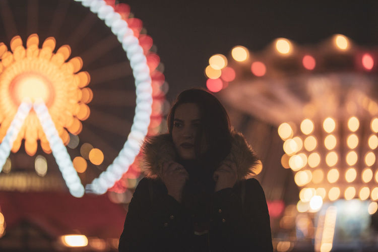 Thoughtful woman standing at illuminated amusement park during night
