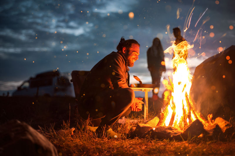 Side view of man crouching by campfire at night