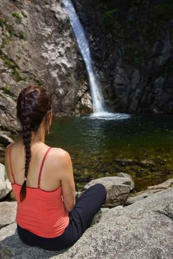 Rear view of woman sitting on rock against waterfall