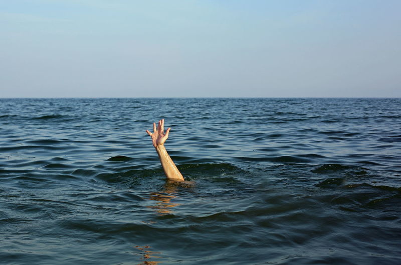 Cropped hand of man coming through sea water against sky