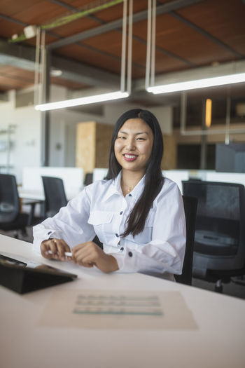 Positive asian female employee sitting at table in workspace and looking at camera