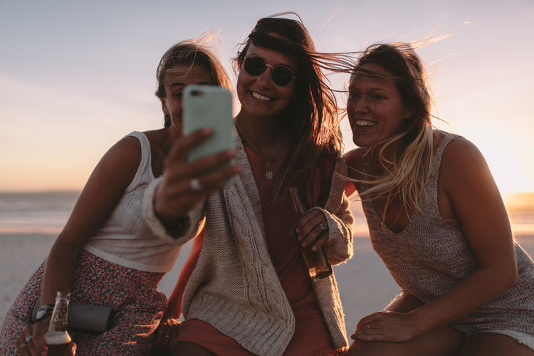 Smiling young woman taking selfie with friends at beach during sunset