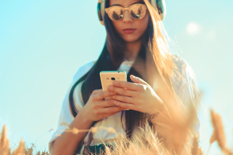 Woman listening music while using mobile phone against clear sky
