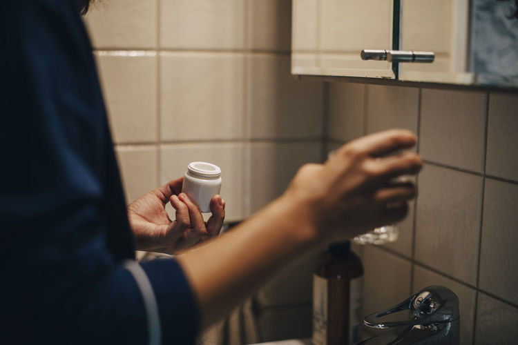 Cropped image of woman holding glass and pills while standing in bathroom