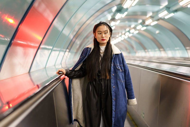 Stylish chinese girl in trendy street fashion outfit, jeans coat, leather beret on subway escalator