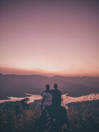 Rear view of couple sitting on mountain against sky during sunset