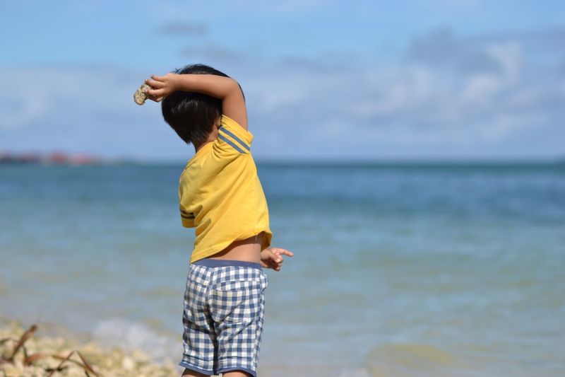 Rear view of boy throwing stone at beach against cloudy sky