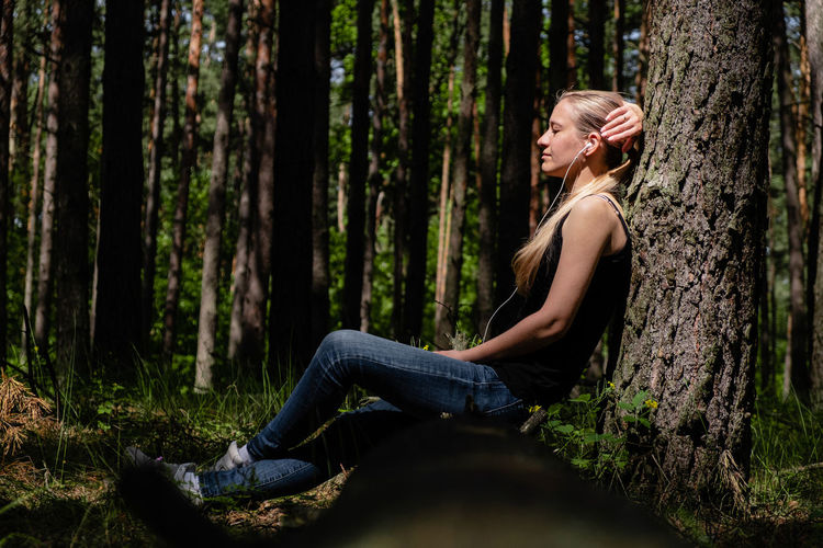 A girl in a forest sits leaning against a tree, with headphones listening to music. digital detox.