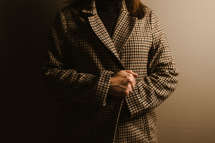 Cropped unrecognizable woman in trendy checkered coat standing against brown wall