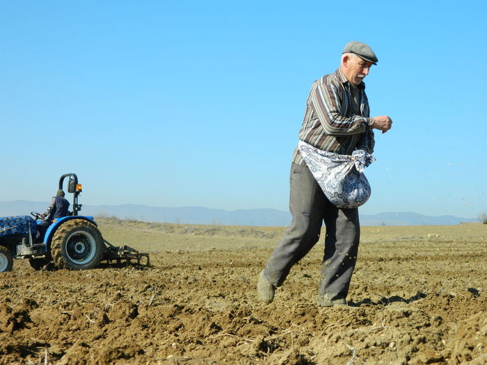 Man scattering seeds with person plowing land with tractor in farm against clear sky