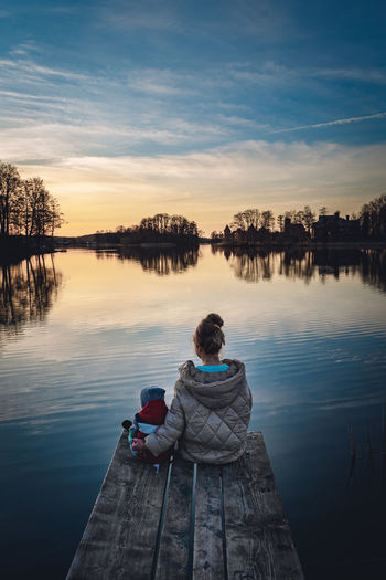 Rear view of mother and son sitting by lake against sky during sunset