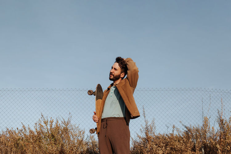 Smiling young bearded male skater in casual wear standing near grass and fence with skateboard and touching hair looking away