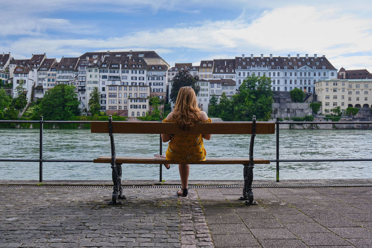 Rear view of woman sitting on bench by river in city