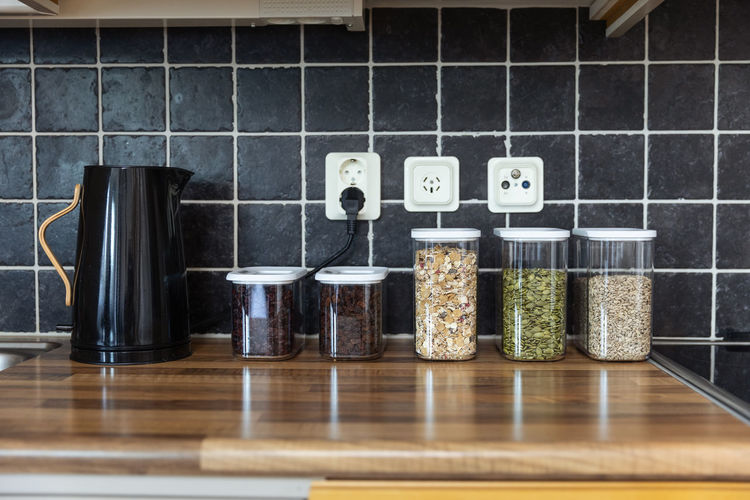 Plastic containers with muesli and sunflower and pumpkin seeds placed near coffee beans and electric kettle on counter in kitchen