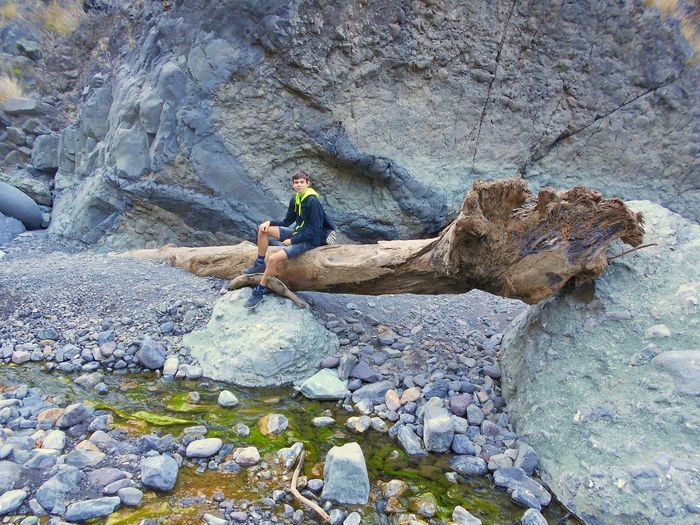 Full length of young man sitting on driftwood by rock formation
