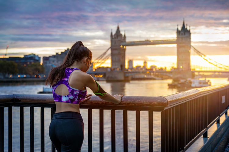 Rear view of woman standing by railing with london bridge in background against sky during sunset