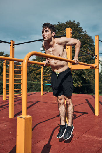 Young shirtless bodybuilder doing dips on parallel bars during his workout in a calisthenics park