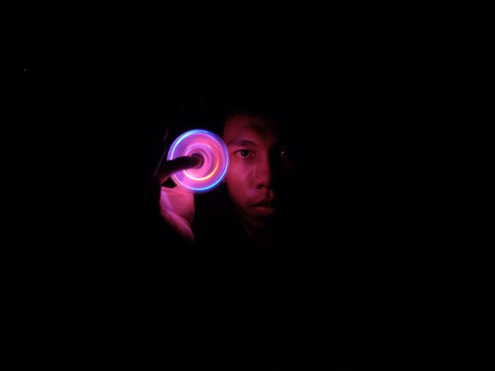 Portrait of young man spinning illuminated fidget spinner against black background