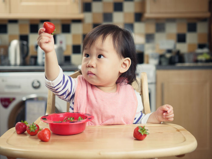 Baby girl with strawberries sitting on high chair at home