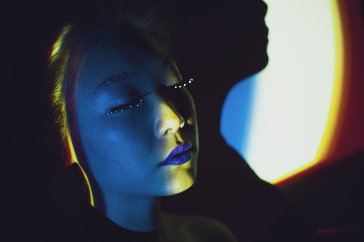 Young asian female model with blond dyed hair and bright makeup with eyes closed standing in dark room illuminated by colorful neon lights